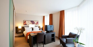 Bright hotel room with double bed, desk, TV, armchairs and large windows with orange curtains.