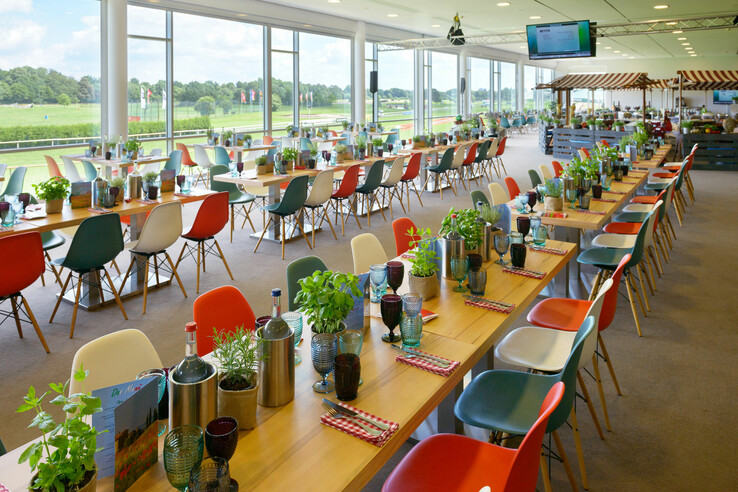 Helles Restaurant mit bunten Stühlen, langen Tischen, Pflanzen und Blick auf die Galopprennbahn durch große Fenster.