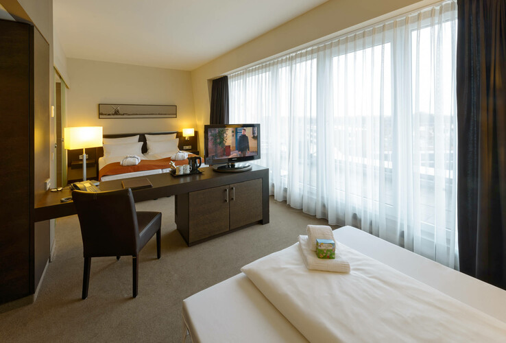 Modernes Hotelzimmer mit Doppelbett, Schreibtisch, TV und großen Fenstern mit Vorhängen im ATLANTIC Hotel Lübeck.