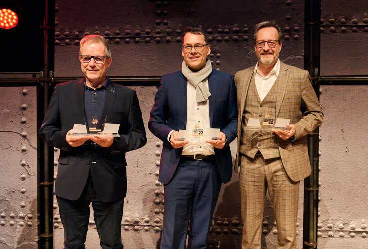 Three men in suits hold awards in front of an industrial wall.