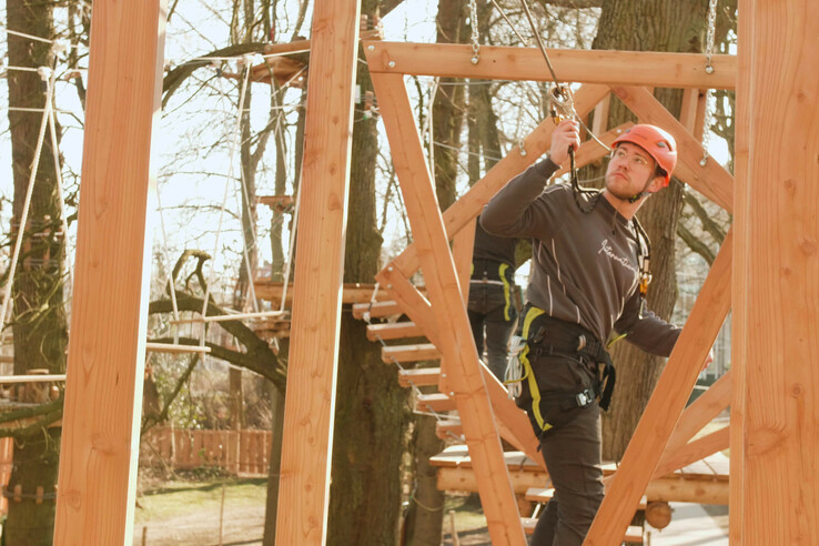 Person im Kletterpark mit Sicherheitsausrüstung und Helm, umgeben von Bäumen und Holzstrukturen.