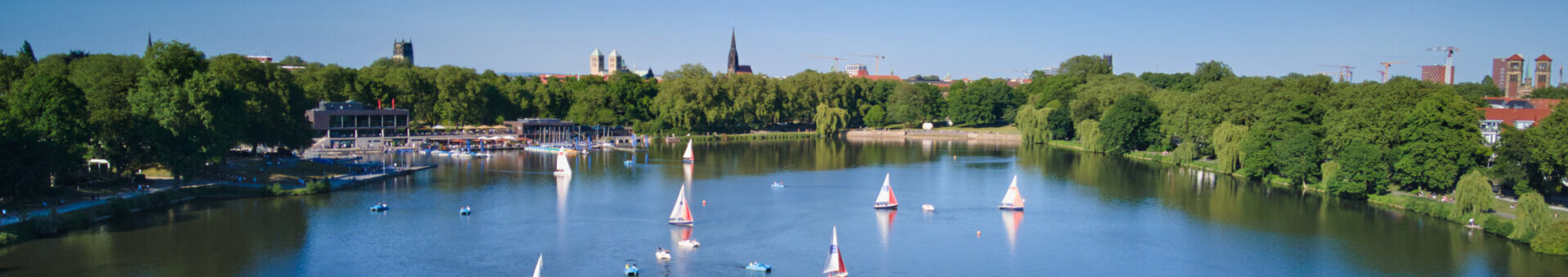 Sailing boats on the Aasee in Münster, surrounded by green trees and blue sky in the background.