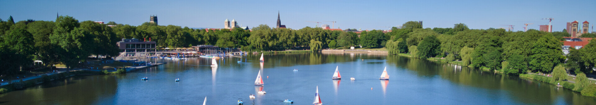 Segelboote auf dem Aasee in Münster, umgeben von grünen Bäumen und blauer Himmel im Hintergrund.
