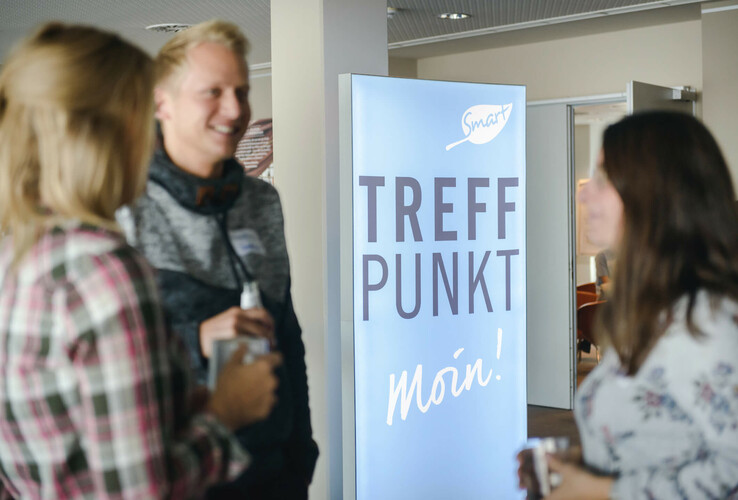 Three people stand in front of an illuminated sign with the inscription "Treffpunkt Moin!" in the ATLANTIC Hotel Sail City.