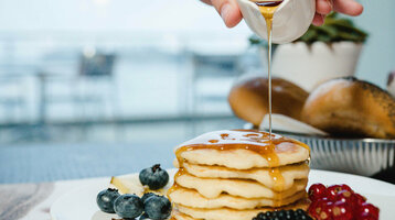 Reichhaltiges Frühstück im ATLANTIC Hotel Wilhelmshaven genießen Ein Stapel Pfannkuchen mit Beeren und Sirup auf einem Teller, im Hintergrund ein unscharfer Tisch.