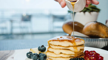 Reichhaltiges Frühstück im ATLANTIC Hotel Wilhelmshaven genießen Ein Stapel Pfannkuchen mit Beeren und Sirup auf einem Teller, im Hintergrund ein unscharfer Tisch.