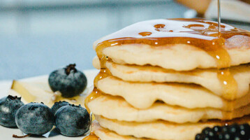 Ein Stapel Pfannkuchen mit Beeren und Sirup auf einem Teller, im Hintergrund ein unscharfer Tisch.