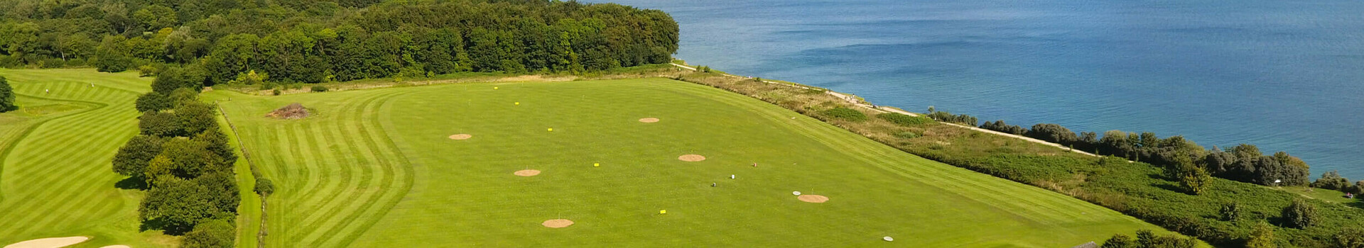 Golf course with sea views, green fairways and sand bunkers, surrounded by trees and a building in the foreground.
