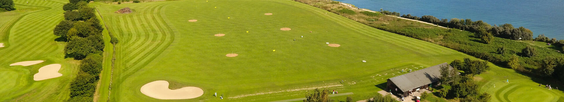 Golfplatz mit Meerblick, grünen Fairways und Sandbunkern, umgeben von Bäumen und einem Gebäude im Vordergrund.