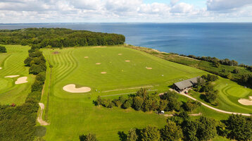 Golfplatz mit Meerblick, grünen Fairways und Sandbunkern, umgeben von Bäumen und einem Gebäude im Vordergrund.