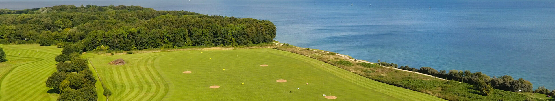 Golf-Arrangement im ATLANTIC Grand Hotel Travemünde Golfplatz mit Meerblick, grünen Fairways und Sandbunkern, umgeben von Bäumen und einem Gebäude im Vordergrund.