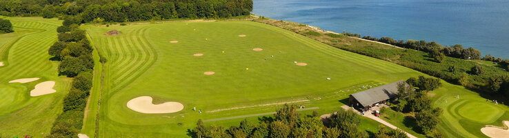 Golf-Arrangement im ATLANTIC Grand Hotel Travemünde Golfplatz mit Meerblick, grünen Fairways und Sandbunkern, umgeben von Bäumen und einem Gebäude im Vordergrund.
