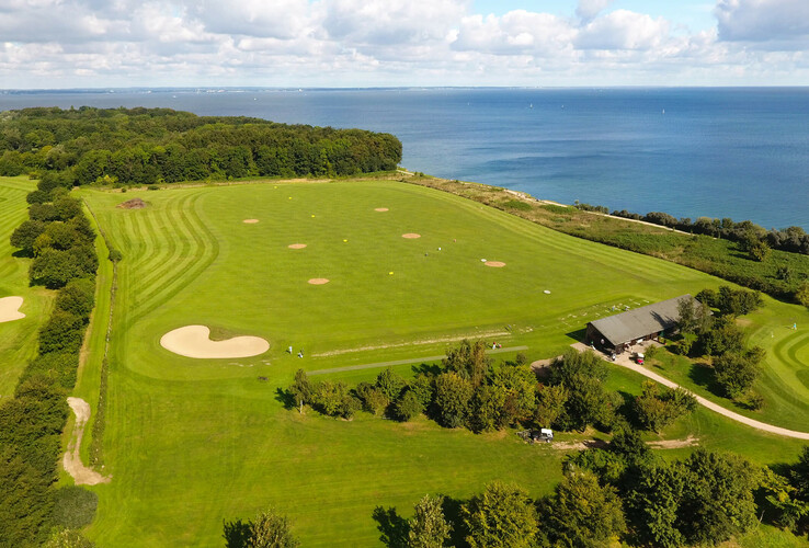 Golf course with sea views, green fairways and sand bunkers, surrounded by trees and a building in the foreground.