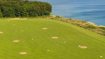 Golf course with sea views, green fairways and sand bunkers, surrounded by trees and a building in the foreground.