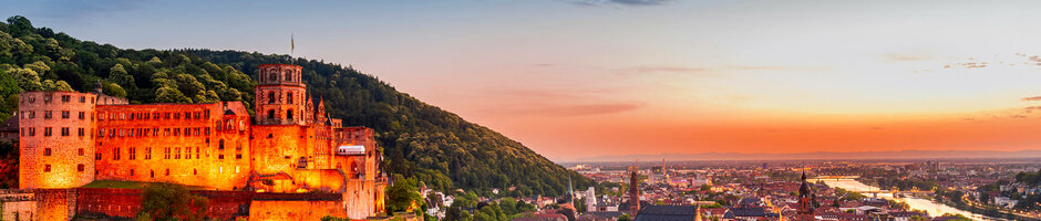 ATLANTIC Hotel Heidelberg Rahmenprogramm Schloss Beleuchtetes Schloss auf Hügel, umgeben von grünen Bäumen, mit Blick auf eine Stadt bei Sonnenuntergang.