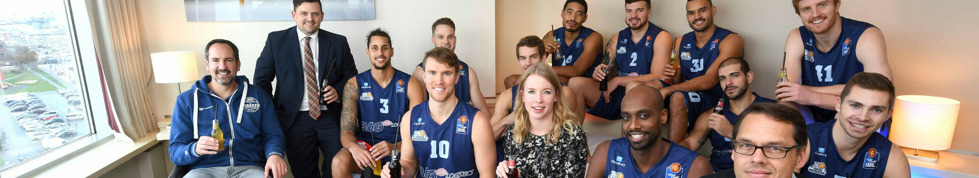ATLANTIC Hotelgruppe bleibt Team Partner der Eisbären Bremerhaven Eine Gruppe von Menschen, darunter Basketballspieler in Trikots, sitzt in einem modernen Hotelzimmer.
