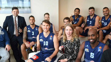 ATLANTIC Hotelgruppe bleibt Team Partner der Eisbären Bremerhaven Eine Gruppe von Menschen, darunter Basketballspieler in Trikots, sitzt in einem modernen Hotelzimmer.