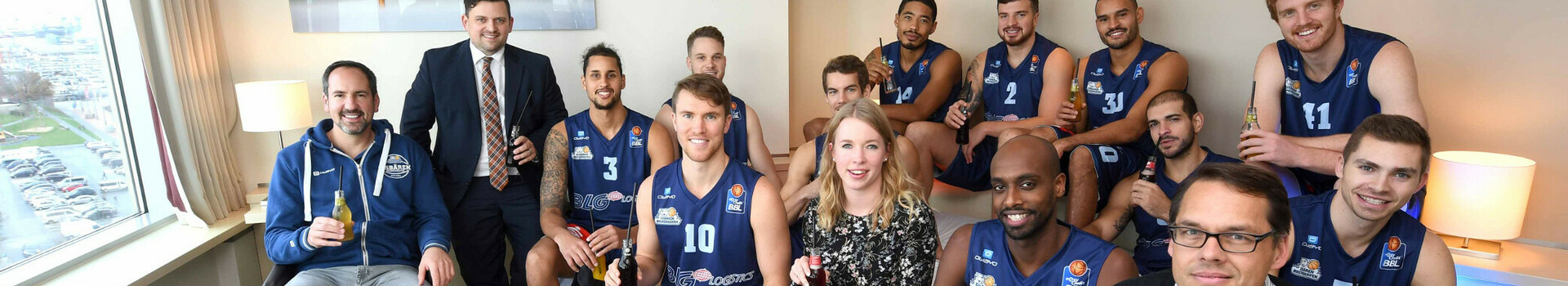 A group of people, including basketball players in jerseys, are sitting in a modern hotel room.