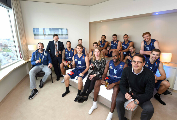 A group of people, including basketball players in jerseys, are sitting in a modern hotel room.