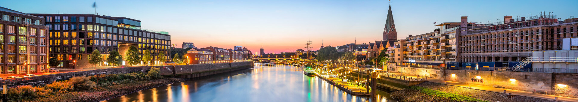 View of the illuminated Weser promenade in Bremen at sunset, with historic buildings and a church tower.