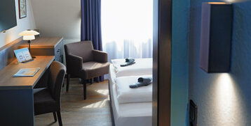 Modernes Hotelzimmer mit Doppelbett, Schreibtisch, braunen Sesseln und Tageslicht durch ein Fenster mit blauen Vorhängen.