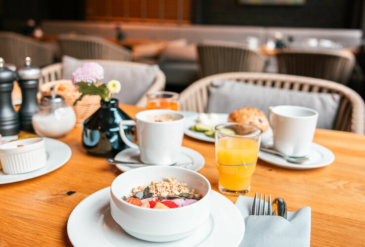 Aufnahme eines Frühstückstisches im Wintergarten. Auf dem Tisch sieht man Gläser mit Saft, verschiedene Tassen Kaffee, einen Teller mit Brötchen und eine Müslischüssel mit Joghurt, Apfelstücken und Knuspermüsli