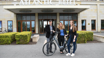 Drei Personen mit Fahrrädern vor dem Eingang des ATLANTIC Hotel Wilhelmshaven.