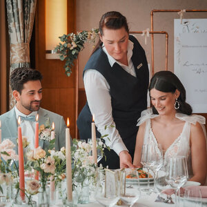 Hochzeitsessen im ATLANTIC Hotel Wilhelmshaven: Ein Paar wird von einer Kellnerin an einem elegant gedeckten Tisch bedient.