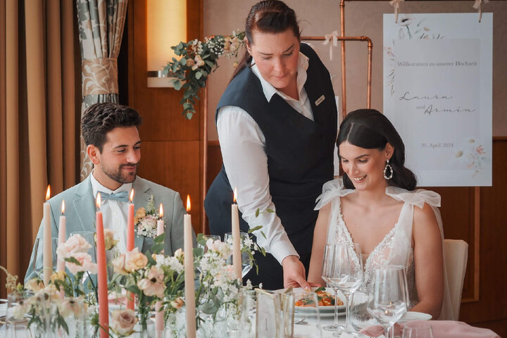 Hochzeitsessen im ATLANTIC Hotel Wilhelmshaven: Ein Paar wird von einer Kellnerin an einem elegant gedeckten Tisch bedient.