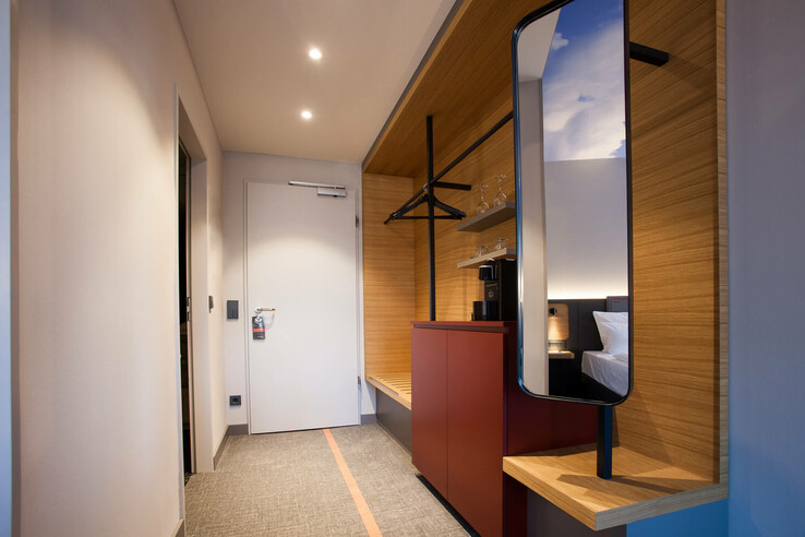 Modern hotel room hallway with wooden details, mirror, clothes rail, wardrobe and view into the bedroom at the ATLANTIC Hotel Airport.
