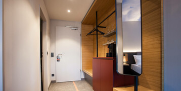 Moderner Hotelzimmerflur mit Holzdetails, Spiegel, Kleiderstange, Schrank und Blick ins Schlafzimmer im ATLANTIC Hotel Airport.