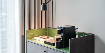 Moderne Kaffeemaschine auf grünem Sideboard, daneben Bücher und Lampe, vor einem Fenster mit Vorhängen im ATLANTIC Hotel Münster.