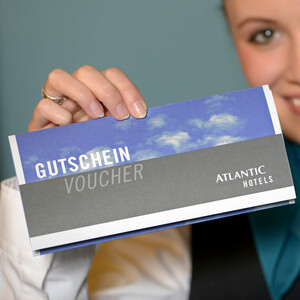 Gutschein-Shop ATLANTIC Hotel Galopprennbahn Eine Hand hält einen Atlantic Hotels Gutschein mit blauem Himmel im Hintergrund.
