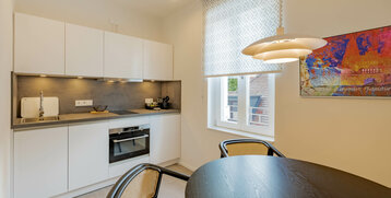 Moderne Hotelküche mit weißen Schränken, Herd, Spüle, rundem Esstisch, Stühlen und Fenster mit gemustertem Rollo.