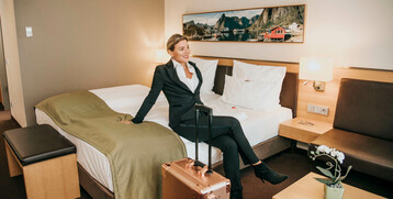Frau in Business-Kleidung sitzt auf Hotelbett, Koffer daneben, modernes Zimmer im ATLANTIC Hotel Kiel.