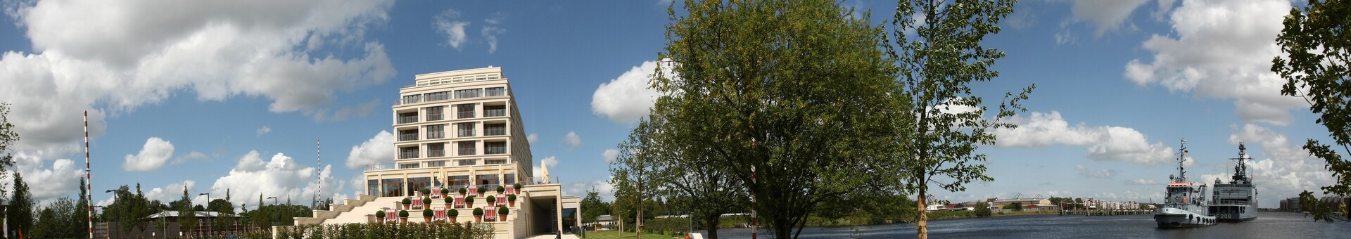 Lage des ATLANTIC Hotel Wilhelmshaven direkt am Ufer des Ems-Jade-Kanal Modernes Hotel am Fluss, umgeben von grüner Wiese und Bäumen, mit Blick auf vorbeifahrende Schiffe.