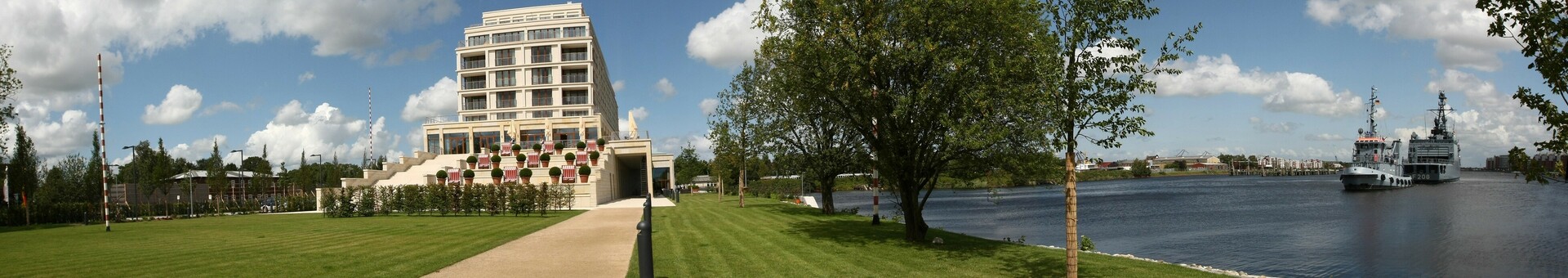 Modernes Hotel am Fluss, umgeben von grüner Wiese und Bäumen, mit Blick auf vorbeifahrende Schiffe.