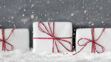 Weiße Geschenke mit roten Schleifen im Schnee vor grauem Hintergrund mit fallenden Schneeflocken.
