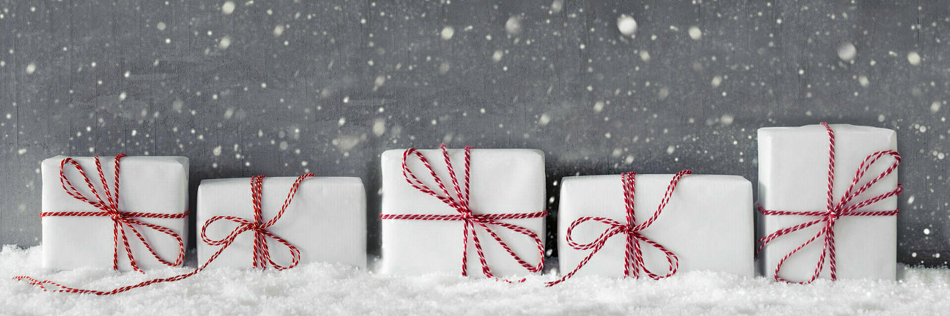 Weiße Geschenke mit roten Schleifen im Schnee vor grauem Hintergrund mit fallenden Schneeflocken.