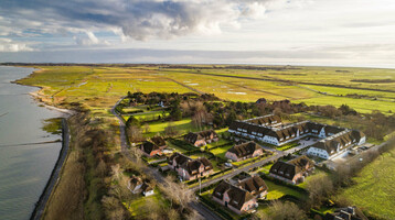 Luftaufnahme eines Küstenhotels mit Reetdachhäusern, umgeben von grünen Feldern und Meerblick unter bewölktem Himmel.