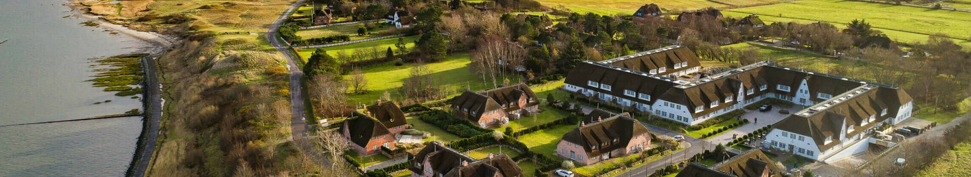 Luftaufnahme eines Küstenhotels mit Reetdachhäusern, umgeben von grünen Feldern und Meerblick unter bewölktem Himmel.