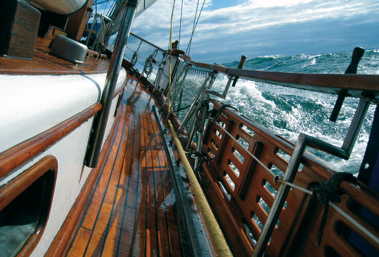 Segelboot auf stürmischer See, Holzdeck glänzt nass, Wellen schlagen gegen Bordwand unter bewölktem Himmel.