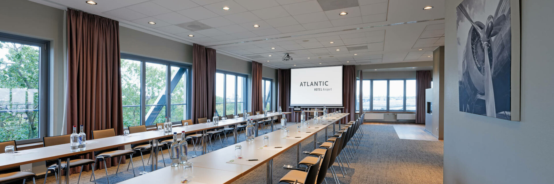 Heller Konferenzraum mit U-förmiger Bestuhlung, großen Fenstern und Projektionsleinwand im ATLANTIC Hotel Airport Bremen.