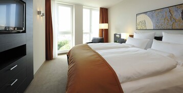 Modernes Hotelzimmer mit großem Bett, Kunst an der Wand, Fernseher, Sessel und bodentiefen Fenstern mit Vorhängen.