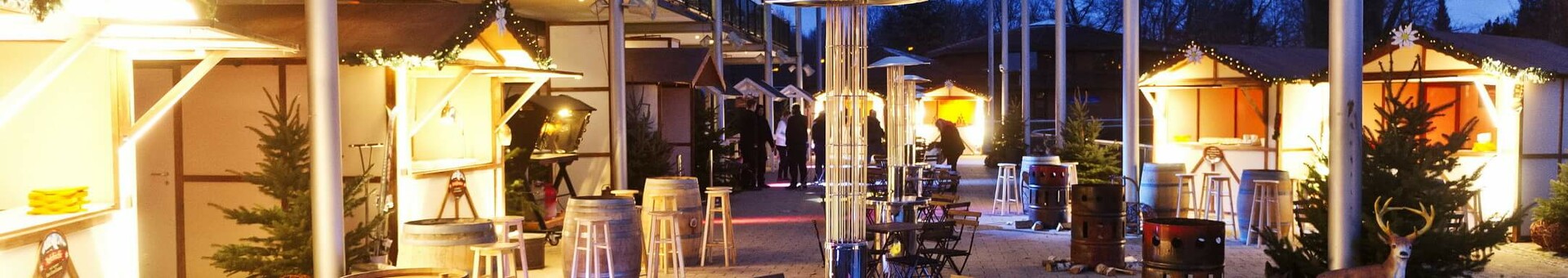 Rahmenprogramm für Weihnachtsfeiern – der kleine Weihnachtsmarkt Beleuchteter Außenbereich mit Holzhütten, Tischen und Stühlen, umgeben von Bäumen und festlicher Dekoration.