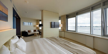 Bright hotel room with large bed, sofa, TV and panoramic windows overlooking the water.