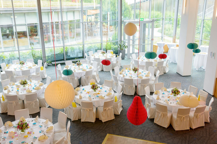 Bright banquet hall with round, white-clothed tables and colorful paper lanterns on the ceiling.