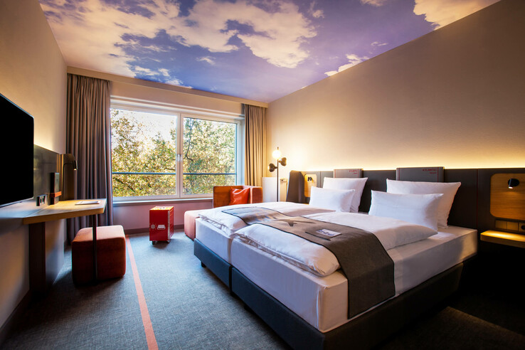 Modern hotel room with double bed, desk, large window and ceiling with cloud motif at the ATLANTIC Hotel Airport Bremen.