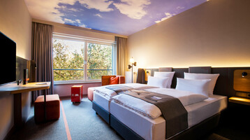 Modern hotel room with double bed, desk, large window and ceiling with cloud motif at the ATLANTIC Hotel Airport Bremen.