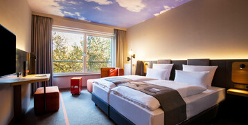 Modernes Hotelzimmer mit Doppelbett, Schreibtisch, großem Fenster und Decke mit Wolkenmotiv im ATLANTIC Hotel Airport Bremen.